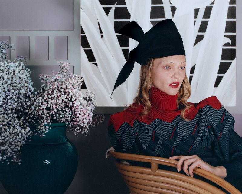 Elegant woman in a unique black hat and patterned dress sits in a rattan chair in an indoor setting with flowers and artwork.