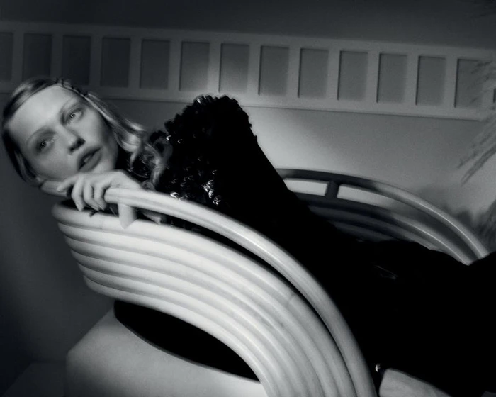 Monochrome portrait of Sasha Pivovarova in a dark embellished top reclining on a curved slatted armchair.