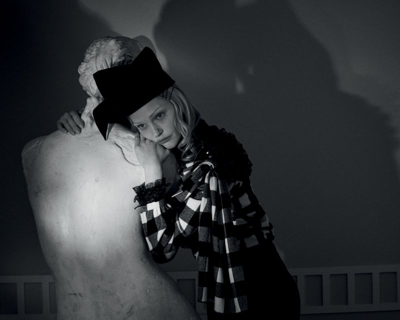 Model Sasha Pivovarova leaning against a white classical statue wearing a black and white patterned top and hat