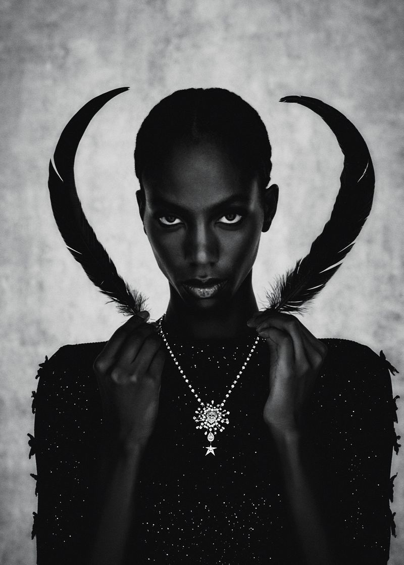 Black and white portrait of an adult woman wearing an embellished top and necklace, holding feathers in a photo studio setting.
