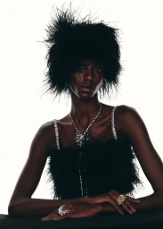 Model Skarla Ali wearing a voluminous black feather headpiece, feathered top, and fine diamond jewelry against a white studio background.