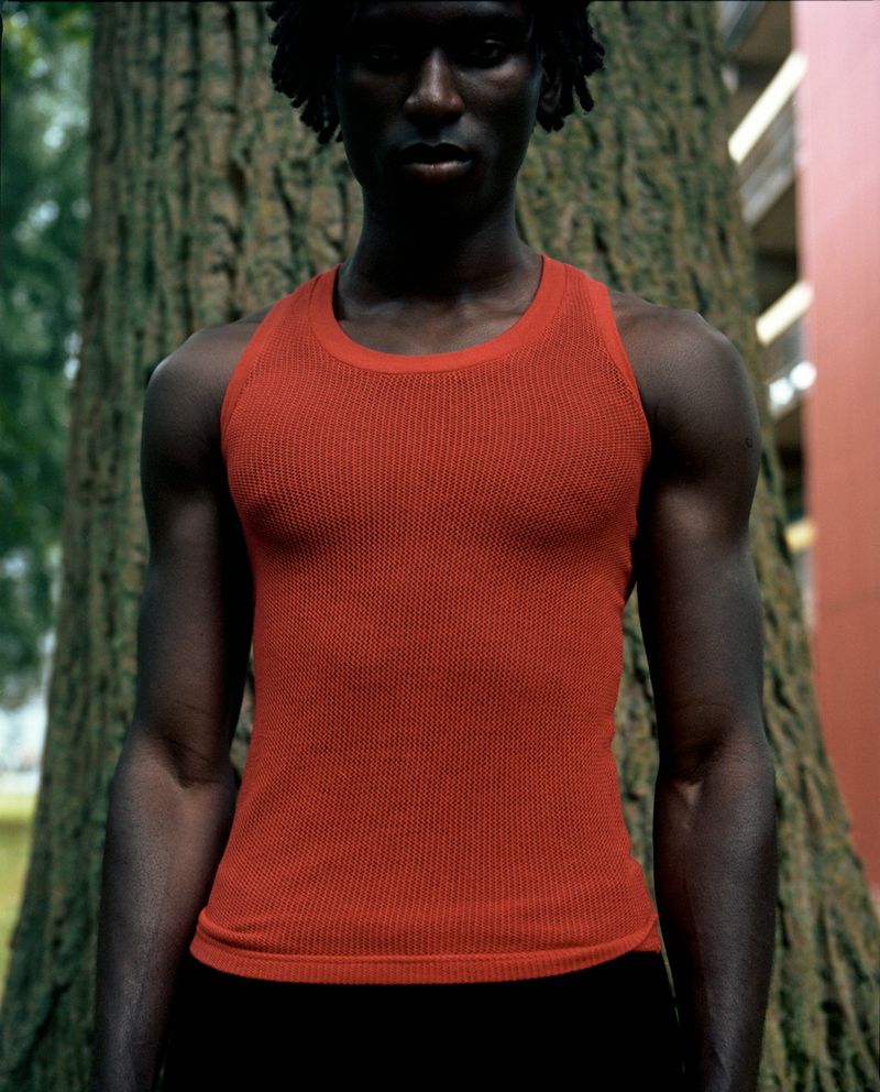Portrait of a Black male wearing a red tank top standing outdoors with a tree trunk in the background and a building in soft focus.