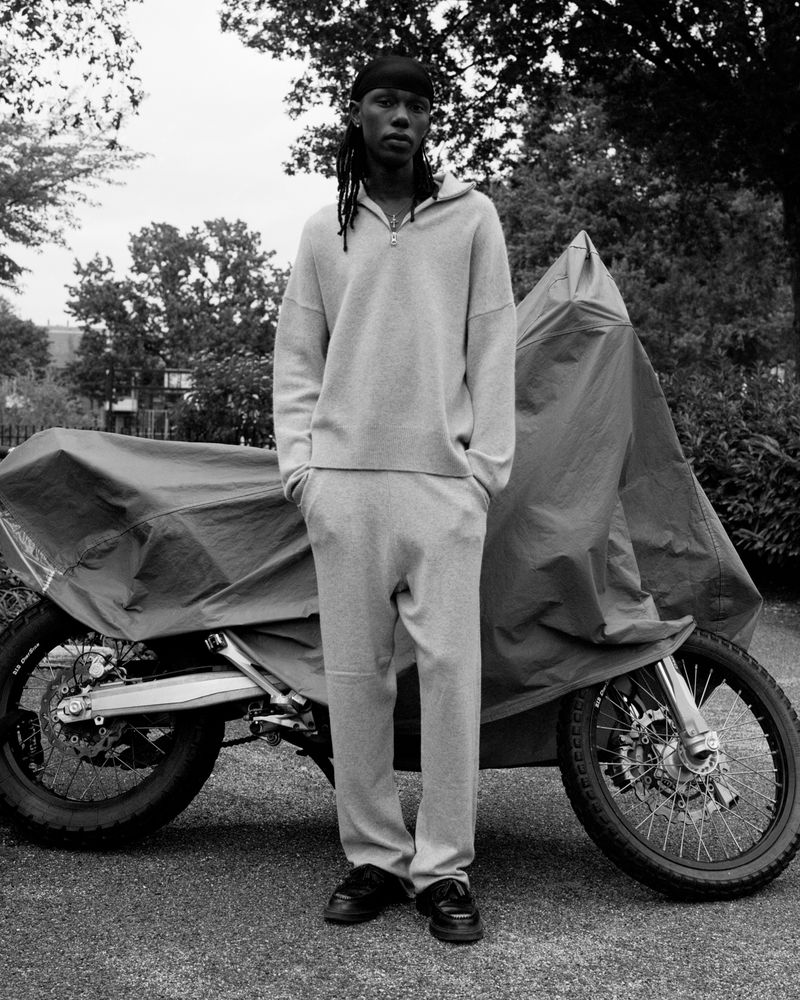 Black and white portrait of a black man in a tracksuit standing in front of a motorcycle covered with a tarp outdoors.