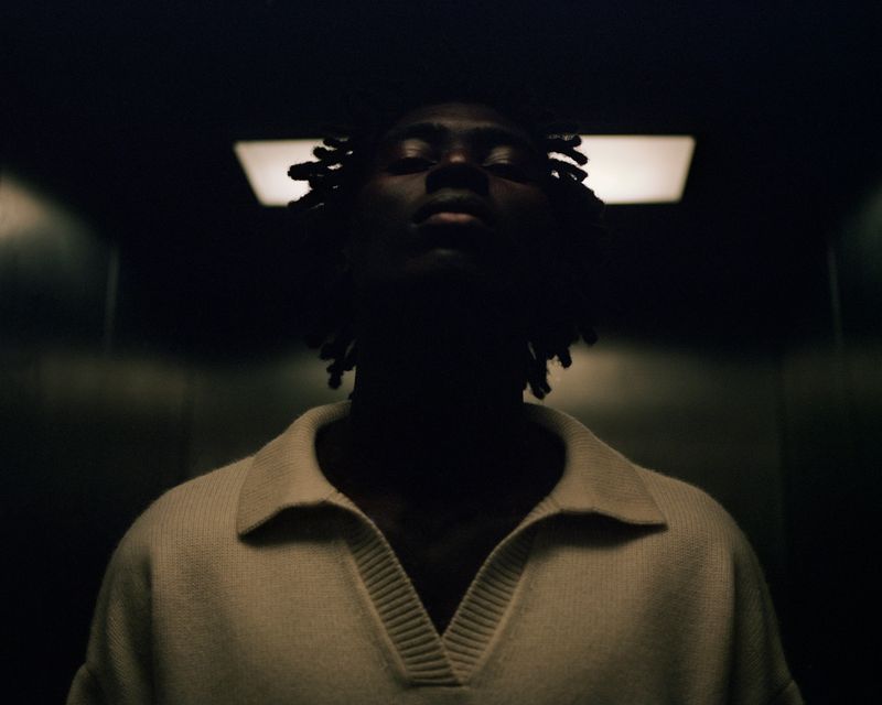 Portrait of African American male with dreadlocks, wearing a white polo shirt, looking up at ceiling light in a dark interior.