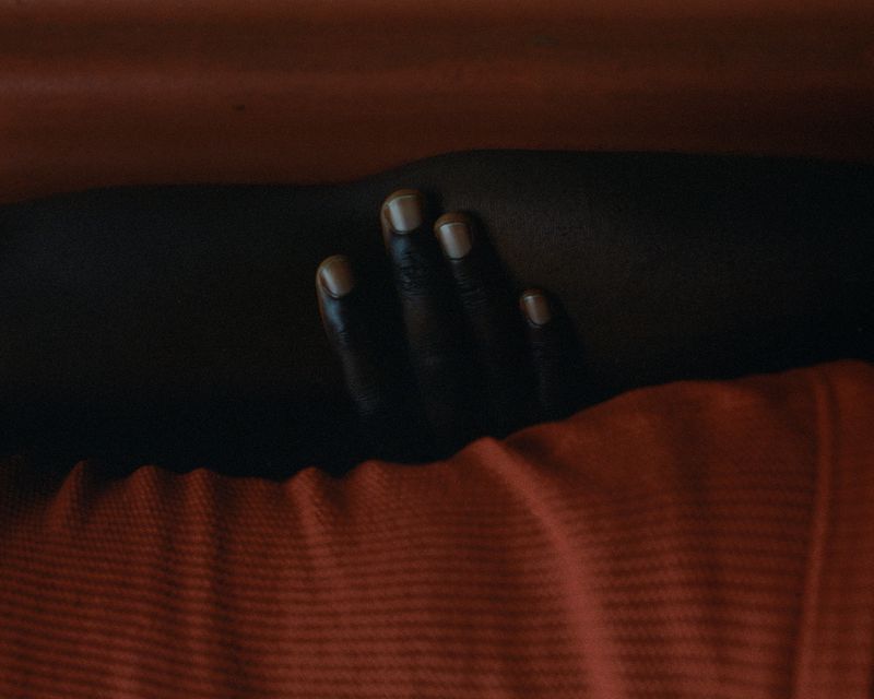 Close-up studio shot of a dark-skinned person's hand with painted nails resting on their arm against a red striped fabric background.