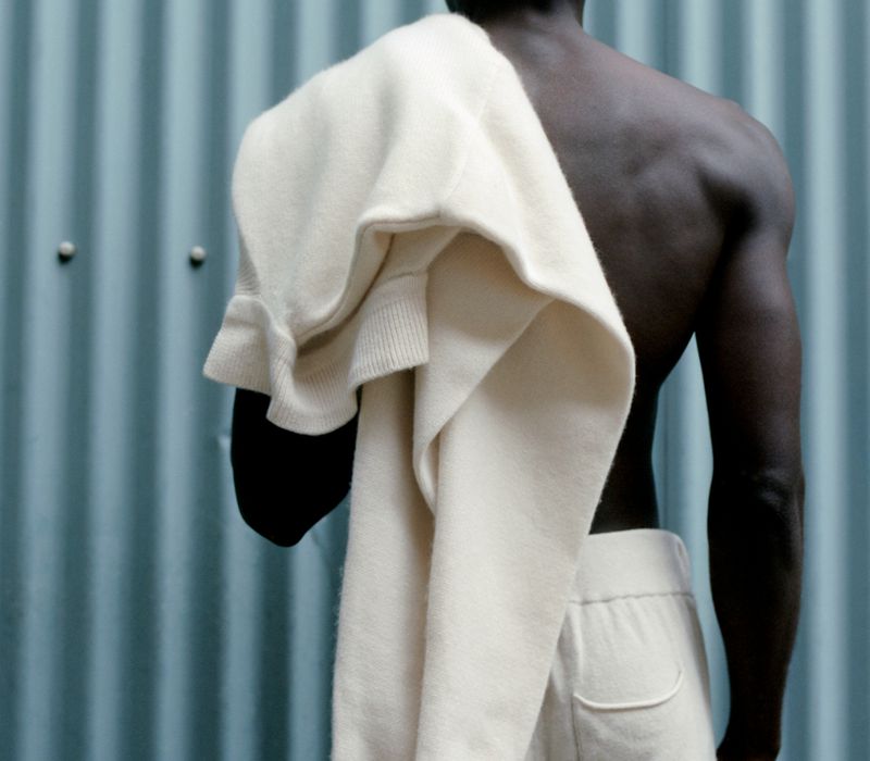 Back view of dark-skinned male with a sweater casually draped over his shoulder, wearing off-white pants against a turquoise wall.
