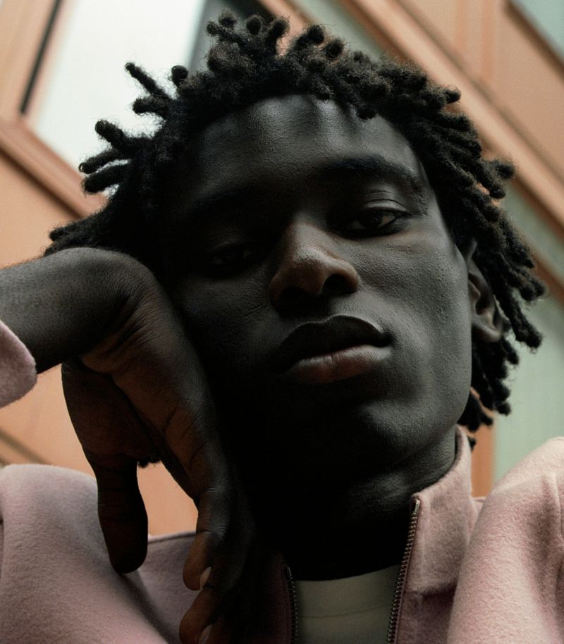 Close-up portrait of an adult male with dreadlocks, wearing a pink jacket, and resting his head on his hand with an architectural background.