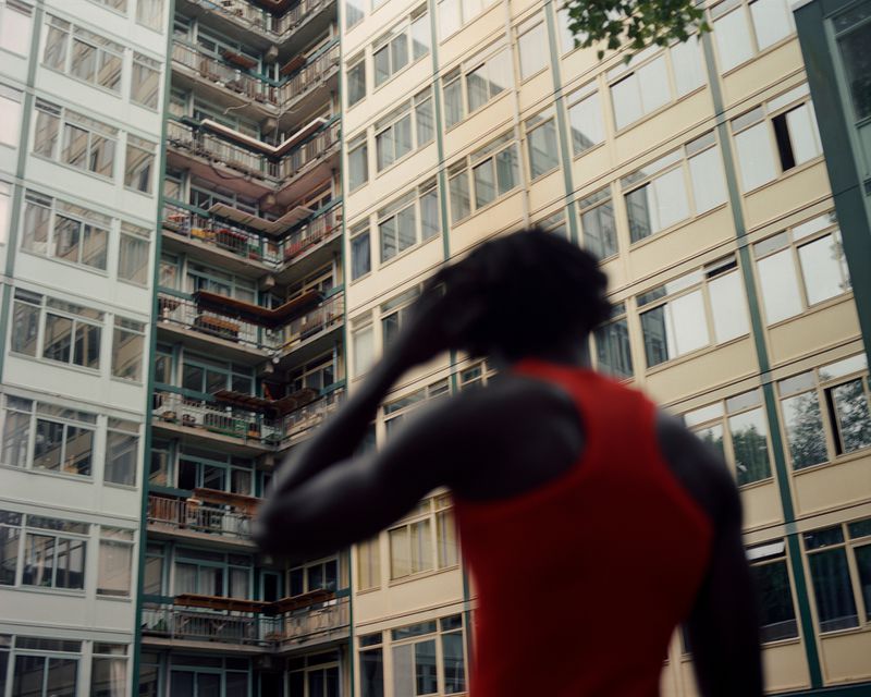 Low-angle view of a Black male walking against a background of a multi-story building with rows of windows and green tree branches.