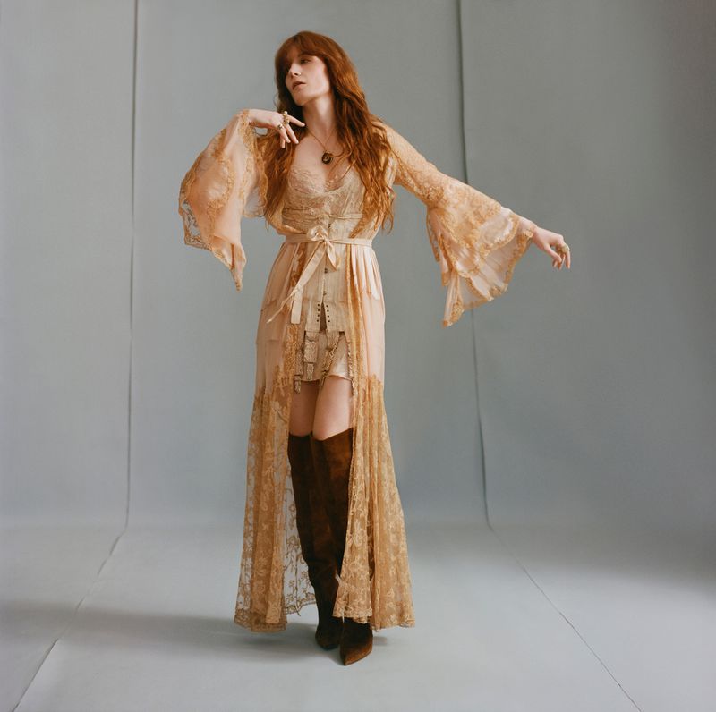 A full shot of a red-haired woman in a lace gown and brown boots striking a pose on a light gray studio background, creating an elegant portrait.