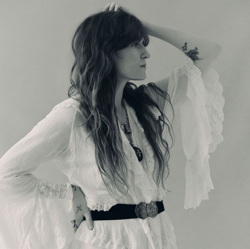 Black and white portrait of a woman with long wavy hair wearing a white ruffled dress and a wide belt in a neutral indoor setting.