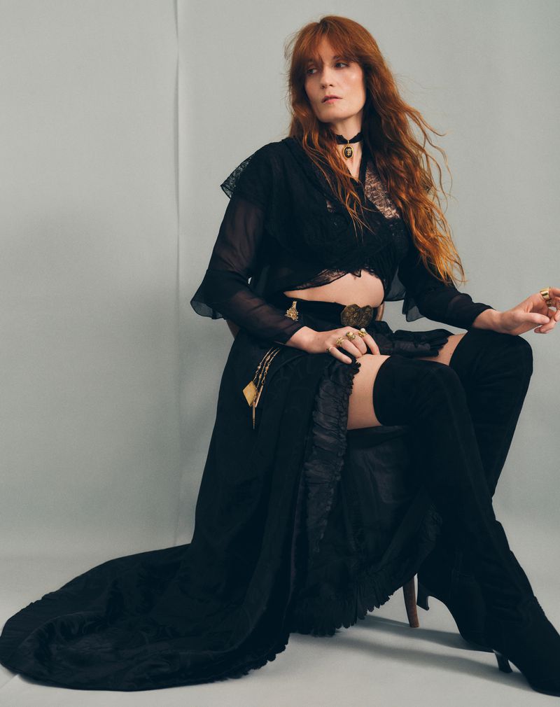 Portrait of an adult female with long red hair sitting in a photo studio wearing a black dress and thigh-high boots against a grey background.