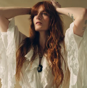 Florence Welch wearing a white lace dress with long ruffled sleeves and a dark pendant necklace against a neutral background.