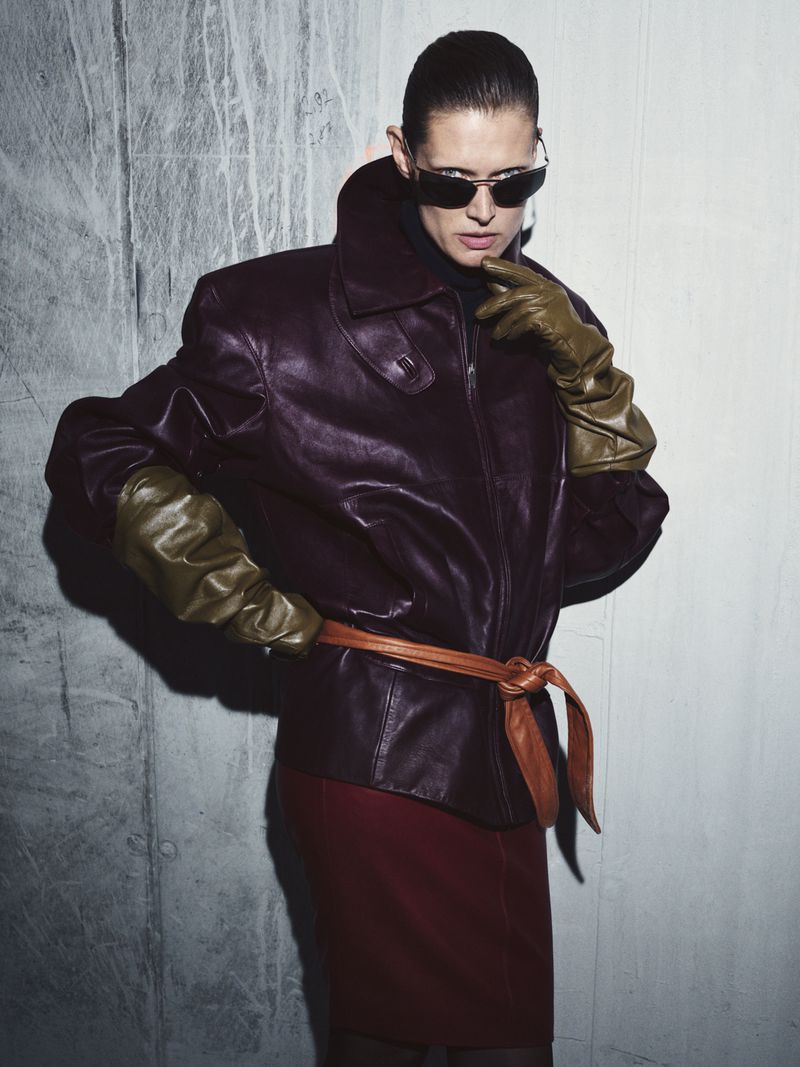 Adult female poses in a studio wearing a leather jacket, gloves, sunglasses, and a red skirt, set against a textured grey background.