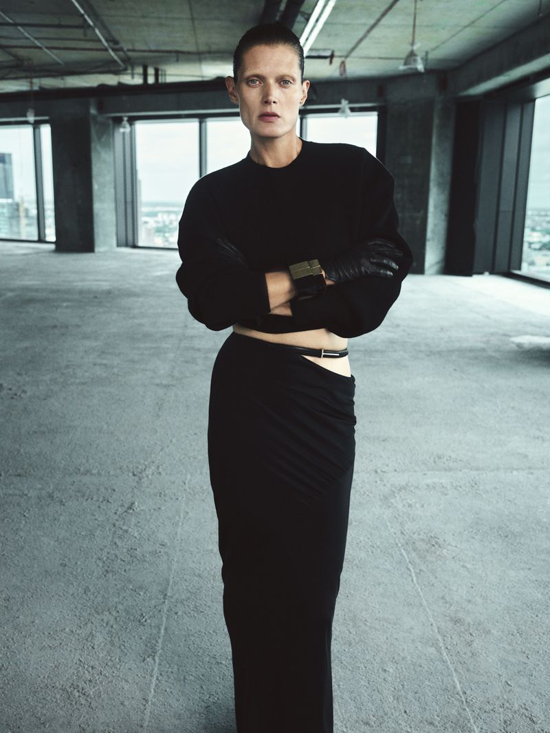 Fashion shot of a woman in a black sweater and black skirt, posing in an empty industrial space, featuring natural light from large windows.