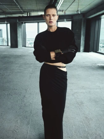 Fashion shot of a woman in a black sweater and black skirt, posing in an empty industrial space, featuring natural light from large windows.
