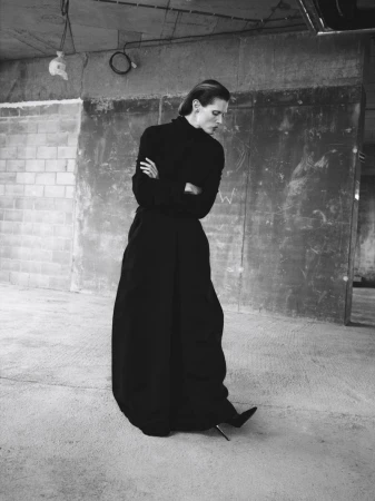 A fashion portrait of an adult woman in a black dress with her arms crossed indoors against a grunge wall in monochrome lighting.