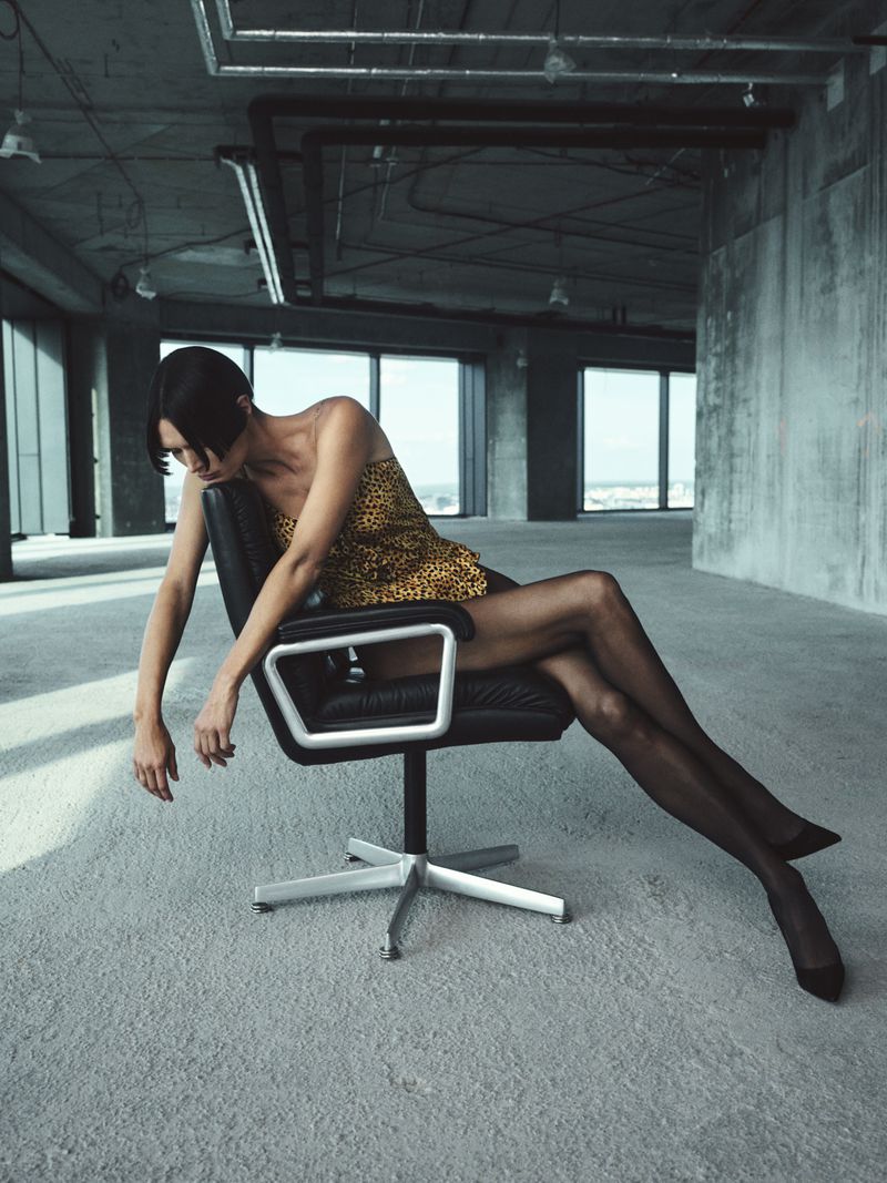 Adult woman sits on an office chair in an empty office space with large windows, wearing a patterned dress and black stockings.