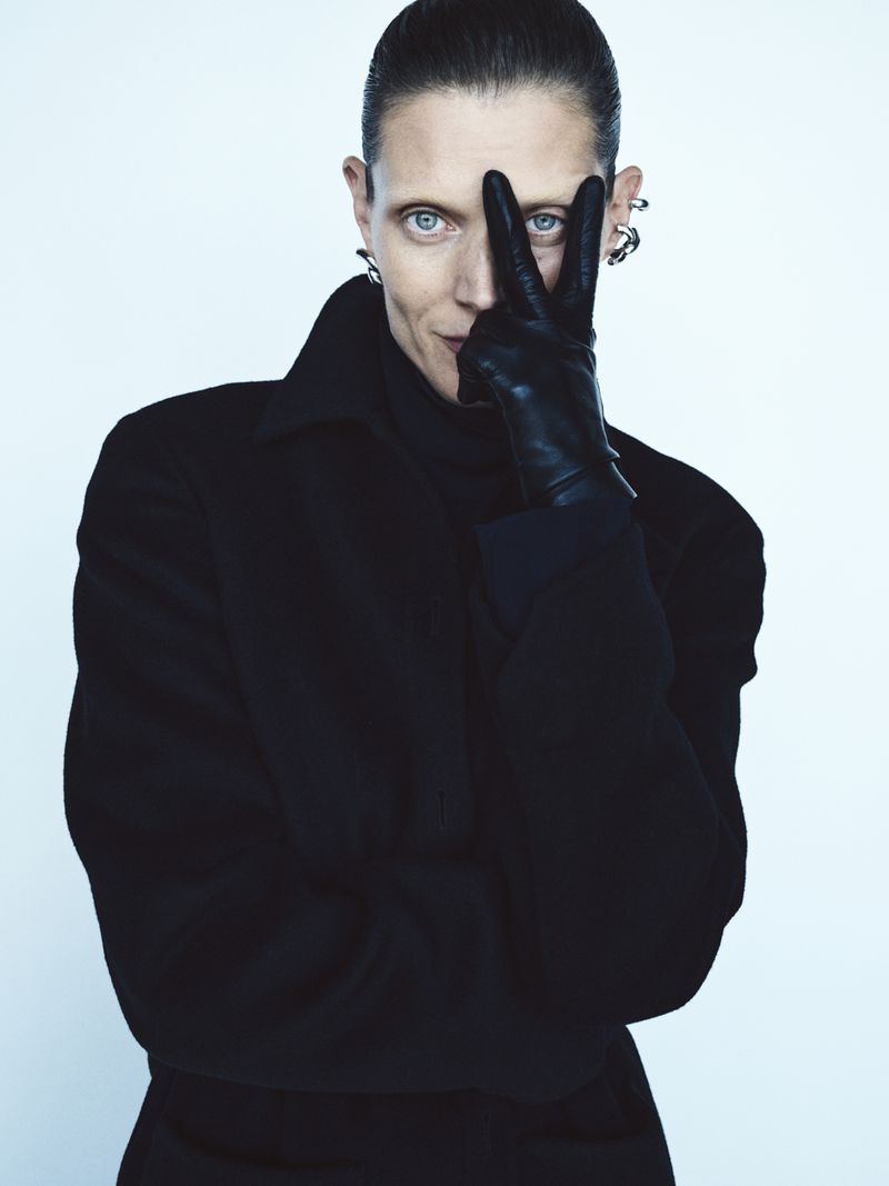 A person wearing a black coat and gloves poses with fingers forming a V shape, set against a bright background in a studio shot.