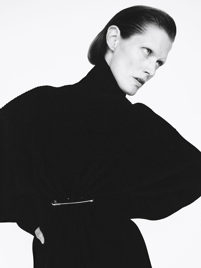 Monochrome fashion portrait of an adult woman with slicked-back hair wearing a black turtleneck sweater with a silver pin in front of a white background.