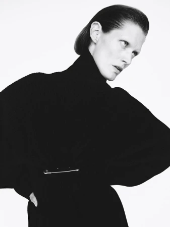 Monochrome fashion portrait of an adult woman with slicked-back hair wearing a black turtleneck sweater with a silver pin in front of a white background.