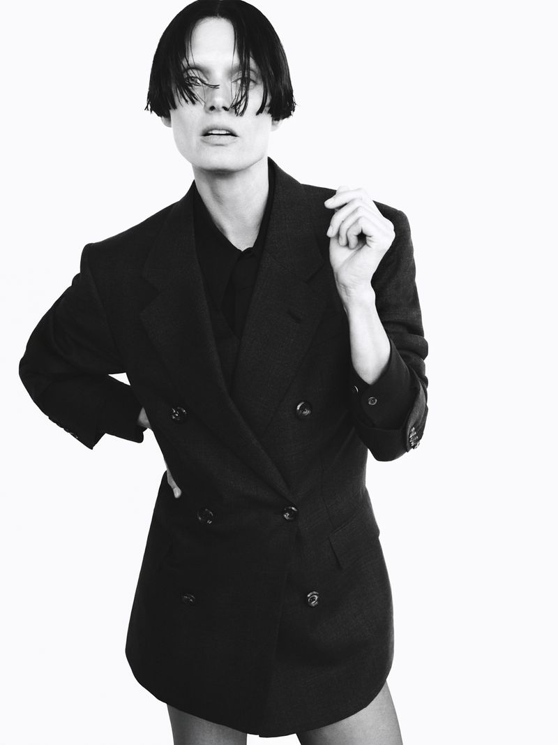 A model poses in a black double-breasted blazer against a white background. The model wears dark hair and has a neutral facial expression.