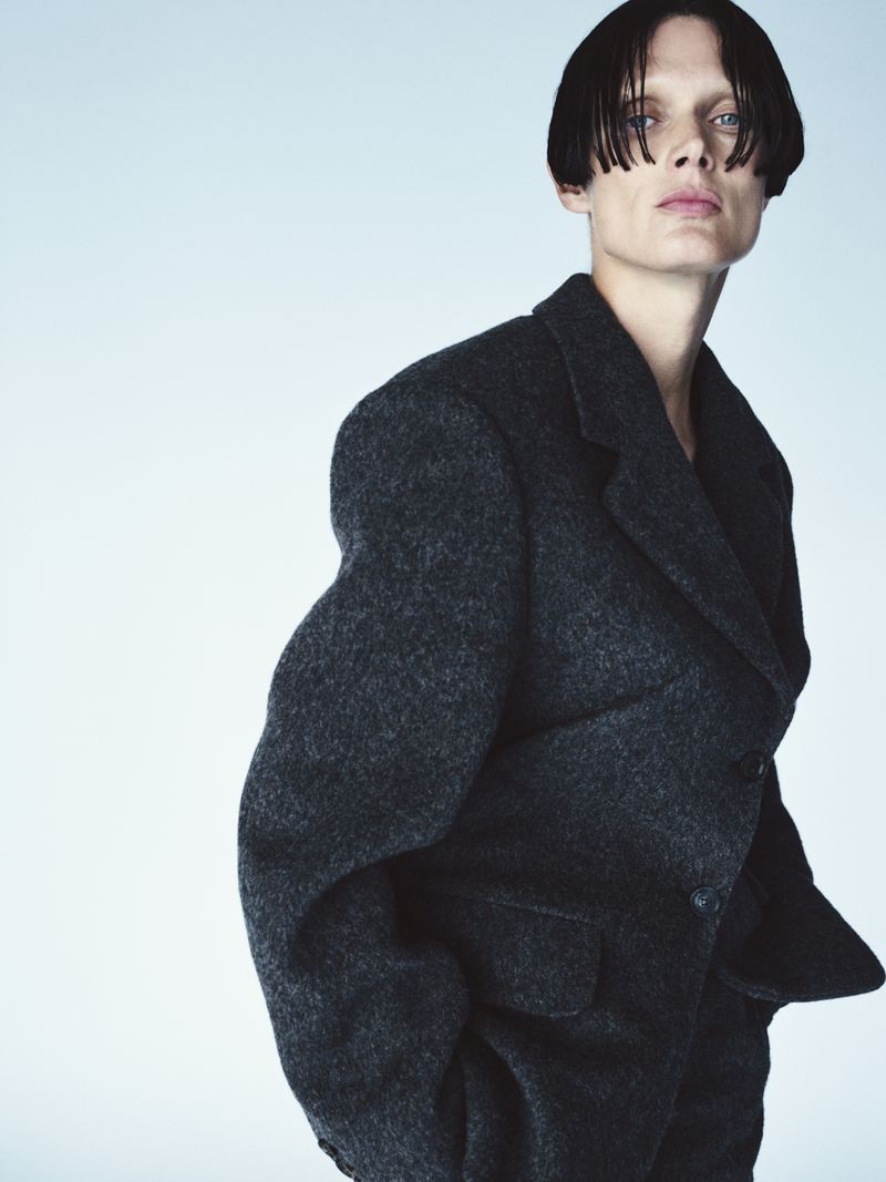 Portrait of a person with short dark hair wearing a dark grey wool coat against a gradient blue background in a studio fashion shoot.