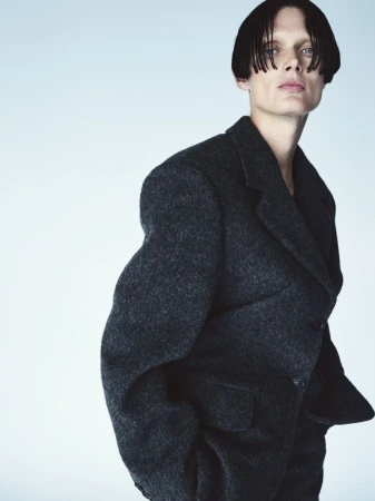 Portrait of a person with short dark hair wearing a dark grey wool coat against a gradient blue background in a studio fashion shoot.