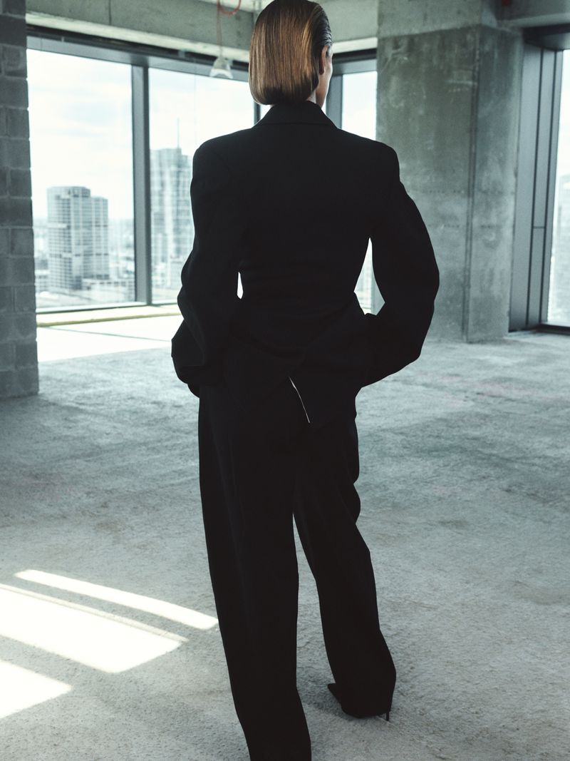 A woman in a black suit stands in an empty room with large windows, overlooking a city skyline, back view, minimalistic architecture.