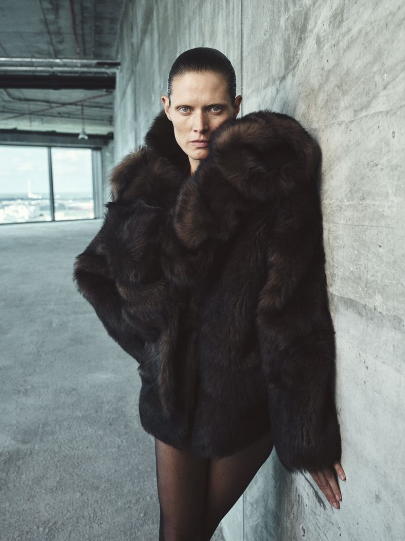 Woman in brown fur coat poses indoors against grey concrete wall with cityscape background. Fashion editorial-style photography.