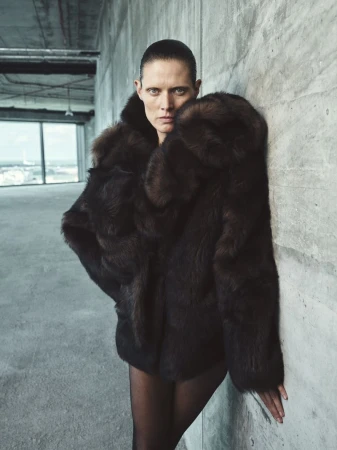 Woman in brown fur coat poses indoors against grey concrete wall with cityscape background. Fashion editorial-style photography.