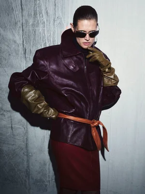 Malgosia Bela wearing an oversized burgundy leather jacket, olive leather gloves, and sunglasses against a textured wall.