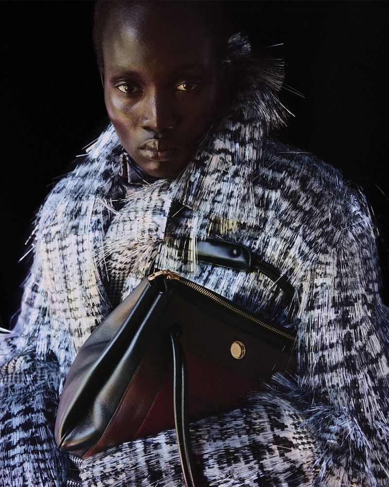 Fashion shot featuring a Black female model in a coat with a textured design, holding a black handbag against a dark background.