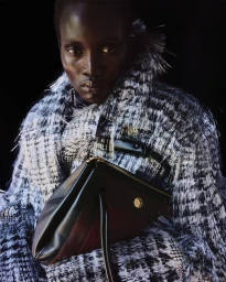 Model Aluel Keror wears a textured fringe coat and holds a black leather handbag against a dark backdrop.