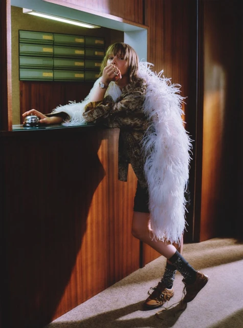 A woman in a patterned jacket and feathered boa leans on a counter next to a bell in an indoor setting with wood paneling.
