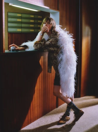 A woman in a patterned jacket and feathered boa leans on a counter next to a bell in an indoor setting with wood paneling.