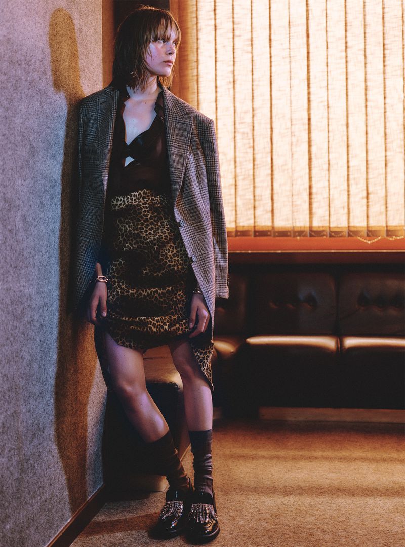 Stylish adult female in leopard print skirt and oversized blazer leans against wall near blinds in a fashion editorial style photograph.