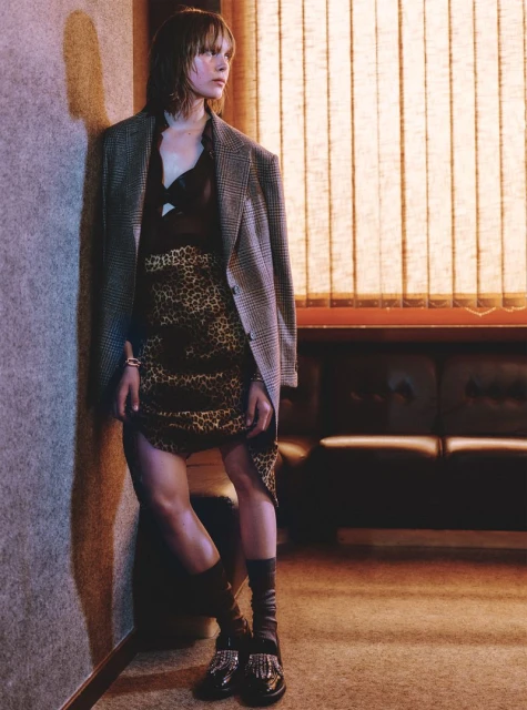 Stylish adult female in leopard print skirt and oversized blazer leans against wall near blinds in a fashion editorial style photograph.