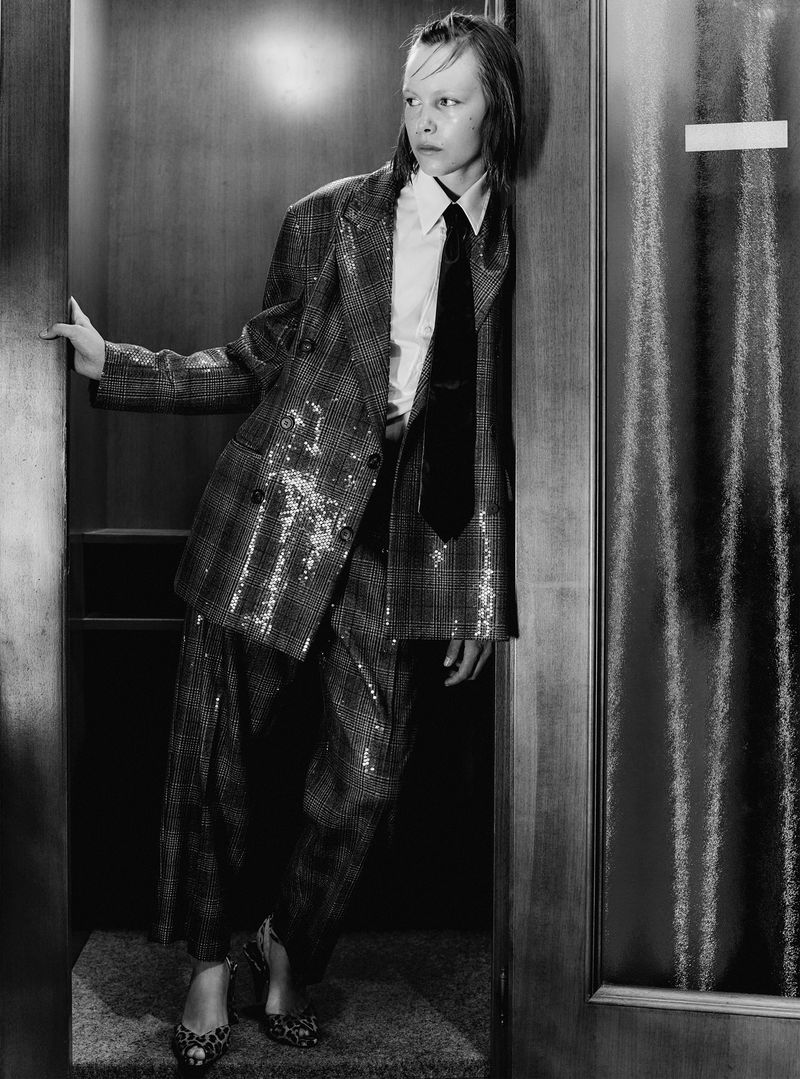 Adult fashion model in a sequined plaid suit and tie leans against a wooden door in a black-and-white fashion editorial shot.