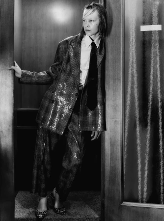 Adult fashion model in a sequined plaid suit and tie leans against a wooden door in a black-and-white fashion editorial shot.