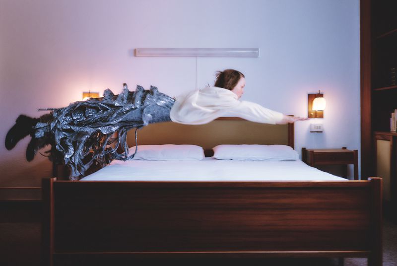 Woman in white poses horizontally on bed with silver sculpture, creates surreal bedroom scene, illuminated by warm lamp light.