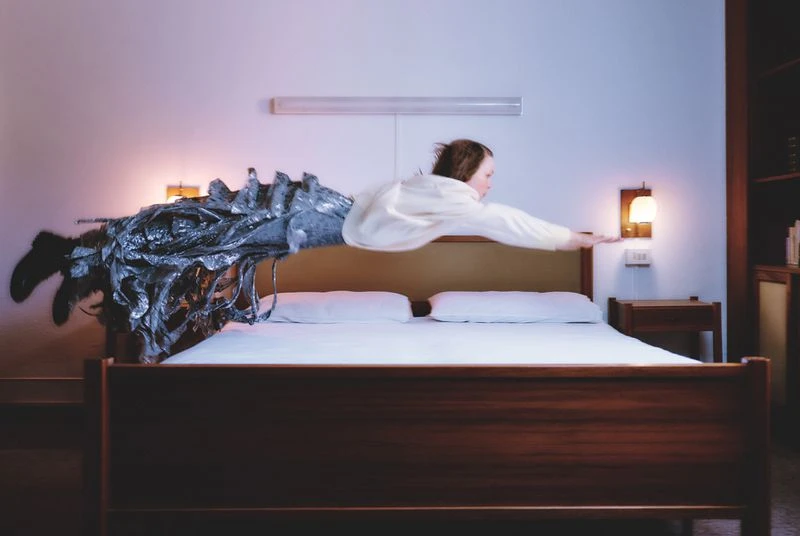 Woman in white poses horizontally on bed with silver sculpture, creates surreal bedroom scene, illuminated by warm lamp light.