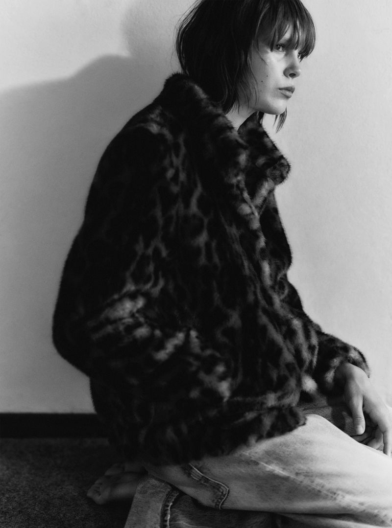 Black and white portrait of a woman in a patterned jacket, kneeing indoors with a neutral background and natural lighting.