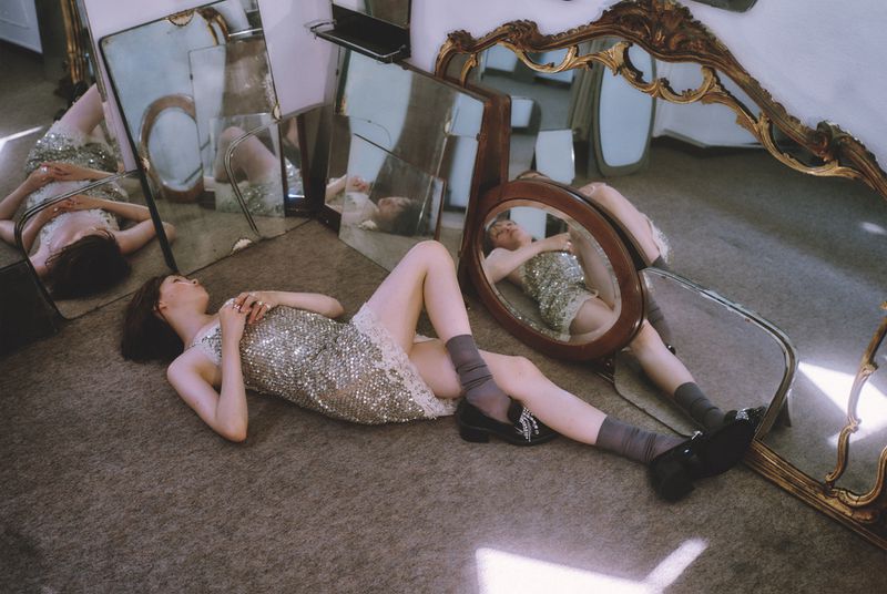 Woman in sparkling dress lies on carpet surrounded by ornate mirrors in a surreal indoor setting with studio lighting and a conceptual theme.