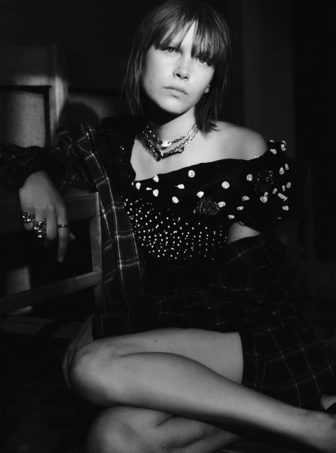 Black and white portrait of a young fair-skinned woman with bangs, wearing an off-the-shoulder top and jewelry, looking at the camera.