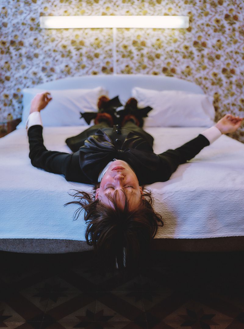 A person in a black suit is lying on a white bed with their arms out to the side, eyes closed, with a floral wall in the background.