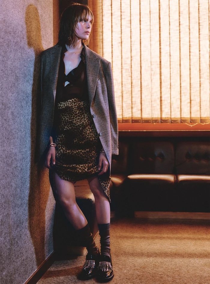 Model Thea Almqvist in an oversized plaid blazer and leopard print skirt leaning against an interior wall.