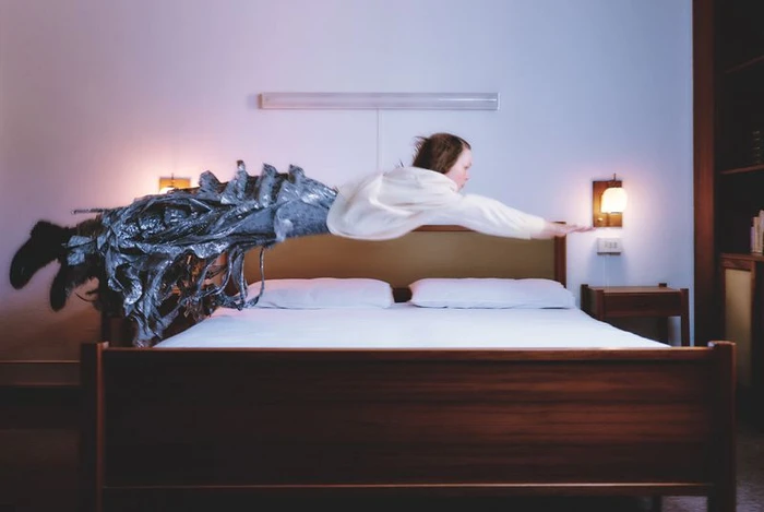 Model Thea Almqvist levitating horizontally above a wooden bed wearing a white top and a textured silver metallic skirt.