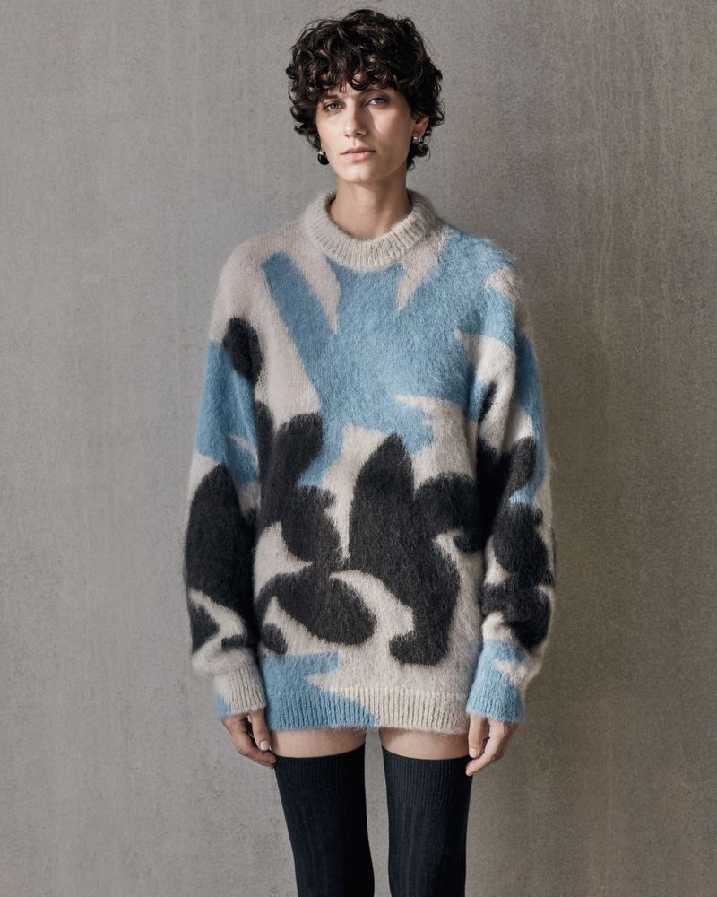 A young woman poses indoors wearing an oversized wool sweater with blue and black abstract patterns, paired with black knee-high socks.
