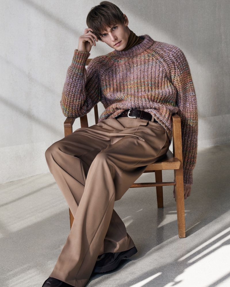 An adult male model with brown hair sits on a wooden chair wearing a multi-colored sweater and taupe pants in a studio setting.