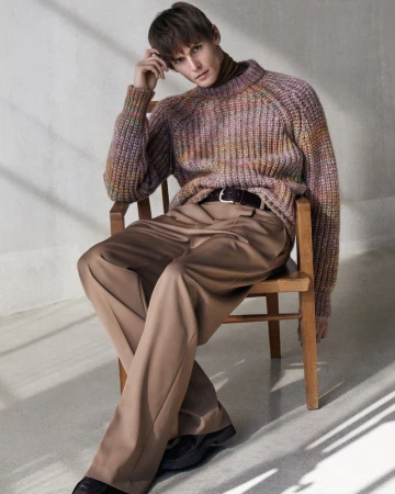 An adult male model with brown hair sits on a wooden chair wearing a multi-colored sweater and taupe pants in a studio setting.